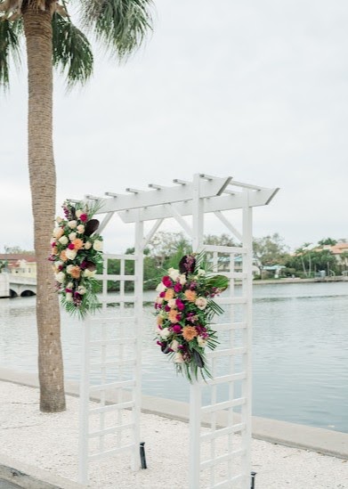 White wedding arbor with tropical floral arrangements beside the water and a palm tree