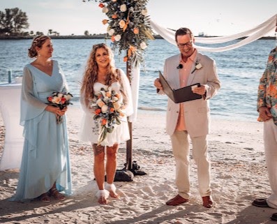 Couple at a beach ceremony with an officiant, beside a peach and cream floral arch