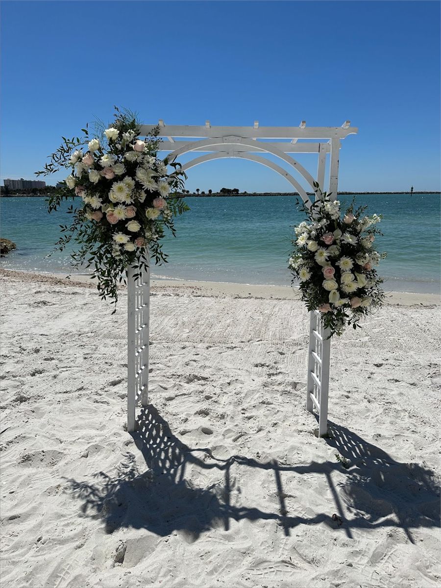 White beach arbor with white and green floral arrangements on the sand beside the water