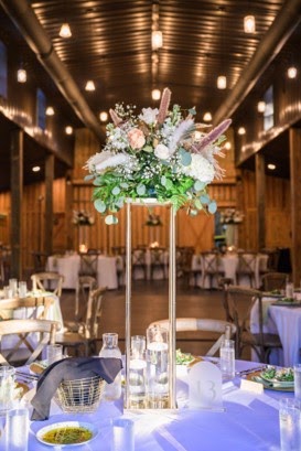Tall centerpiece of blush and white flowers with greenery on a reception table in a wooden barn