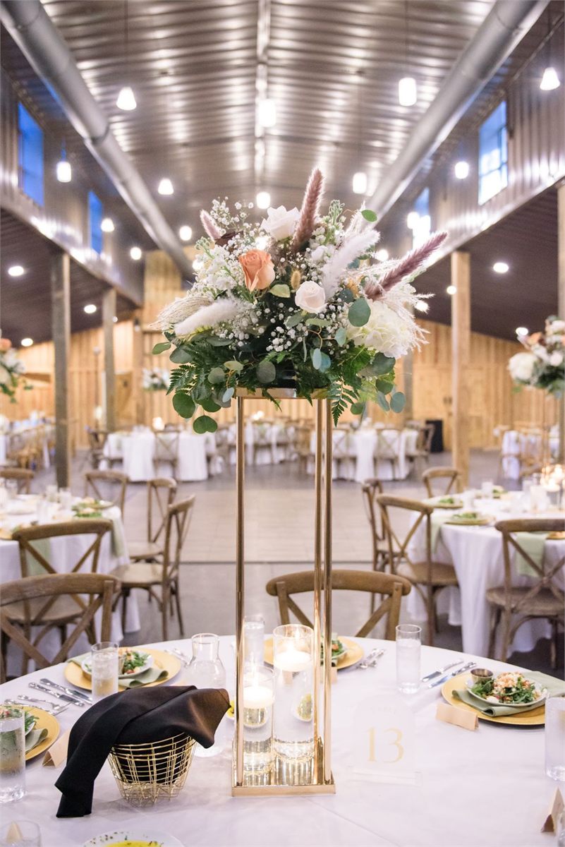 Tall centerpiece of white flowers and dried grasses on a gold stand in a barn reception