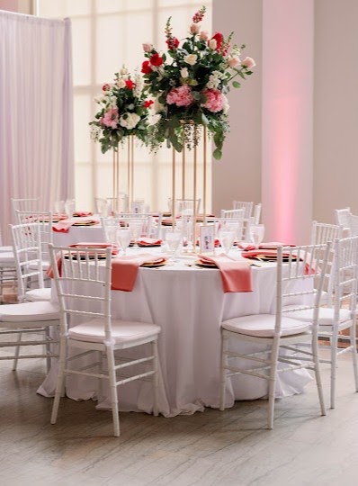 Tall pink and red centerpiece on a gold stand above a round table with white chairs