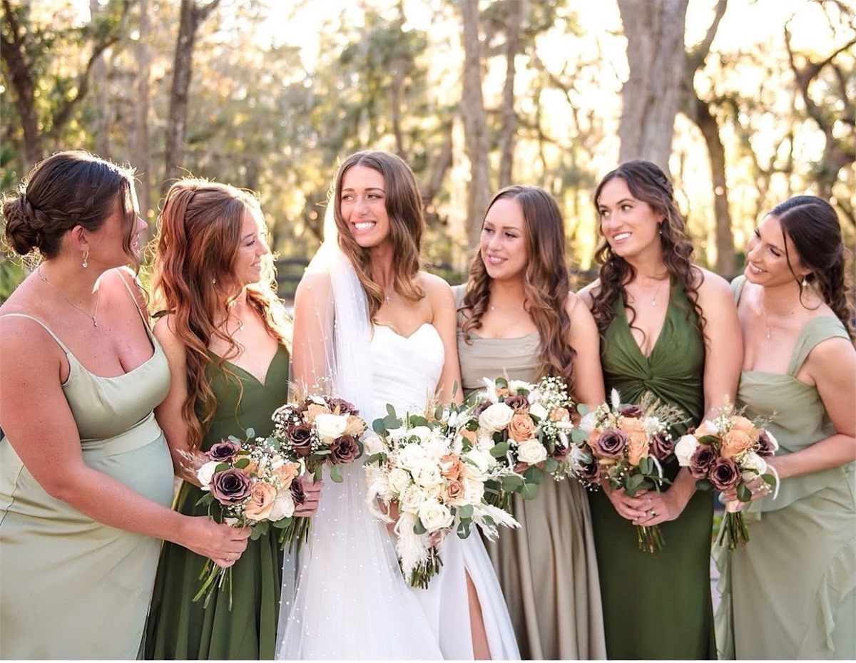 Bride with five bridesmaids in green dresses, each holding a bouquet, outdoors in late afternoon light