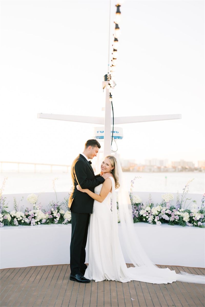 Bride and groom embracing on a dock at sunset beside white floral arrangements