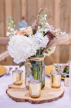 Rustic centerpiece of white hydrangea and dried grasses in a mason jar on a wood slice, with gold votive candles