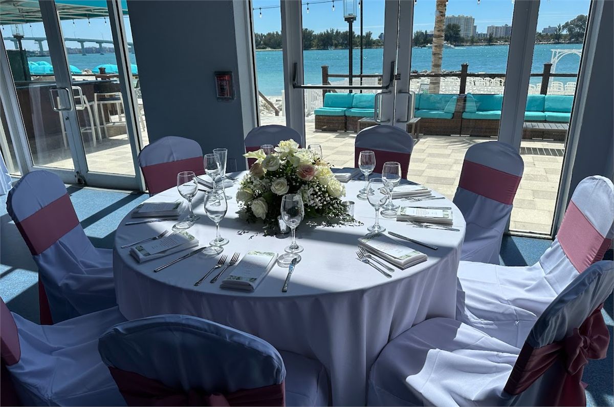 Round reception table with white linens, pink chair sashes and a white centerpiece beside waterfront windows